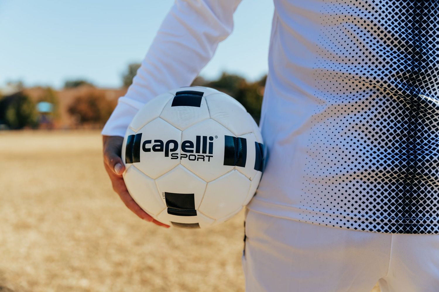 Hand holding a white and black Capelli Sport soccer ball on an outdoor field.