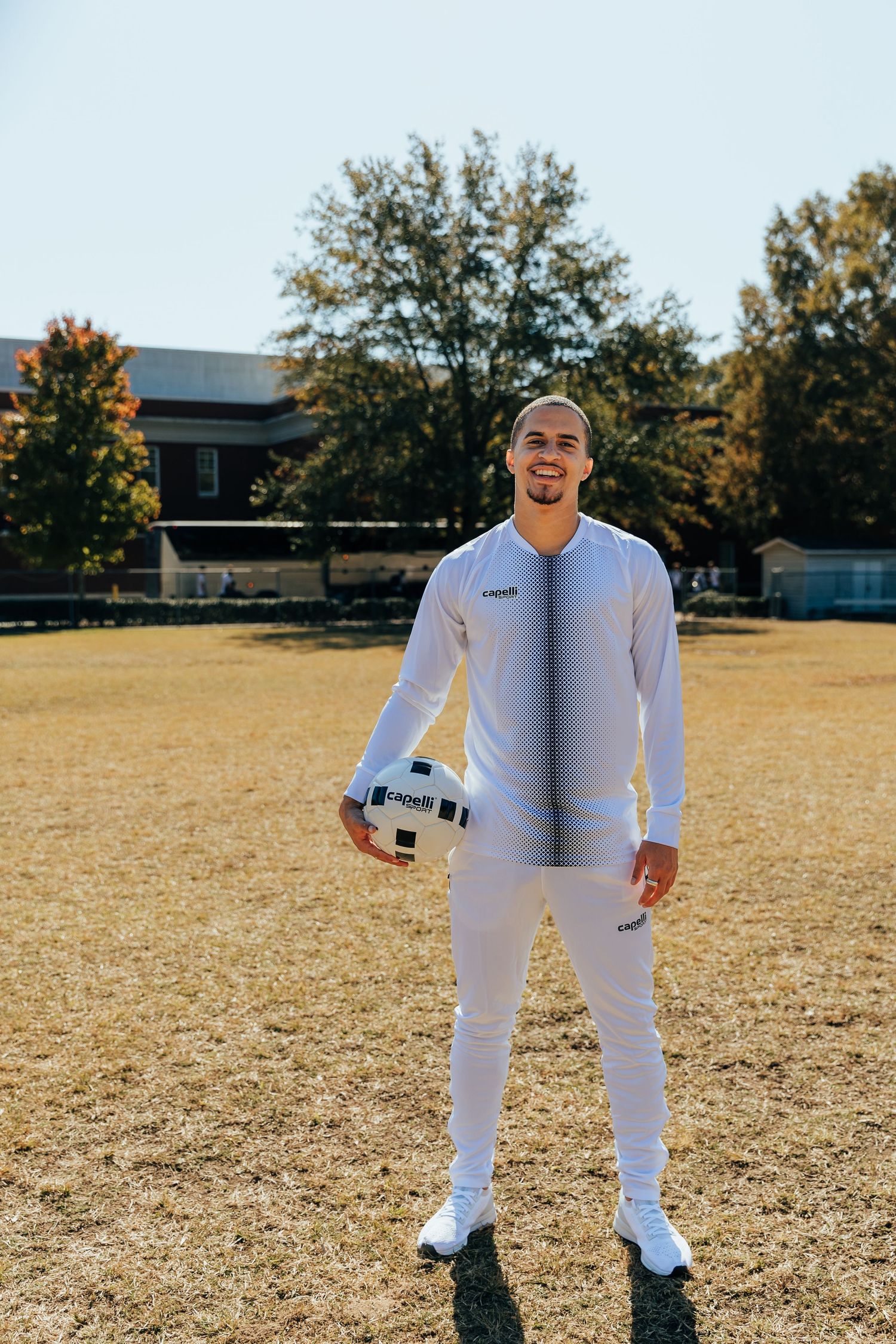 Male athlete in full white Capelli Sport tracksuit smiling while holding a soccer ball on a field.