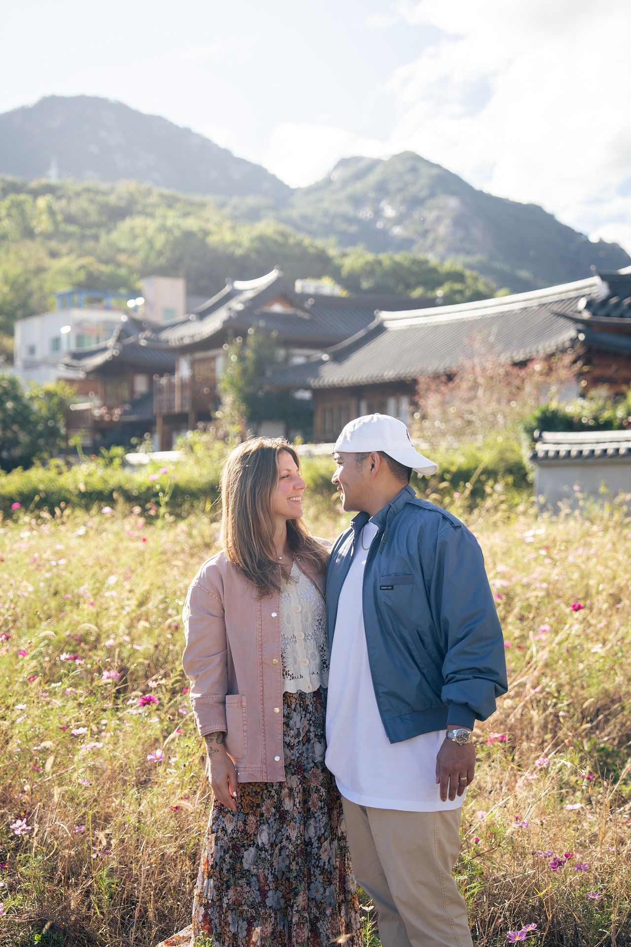 Couple standing together in a sunlit meadow with mountains and traditional Korean architecture in the background.
