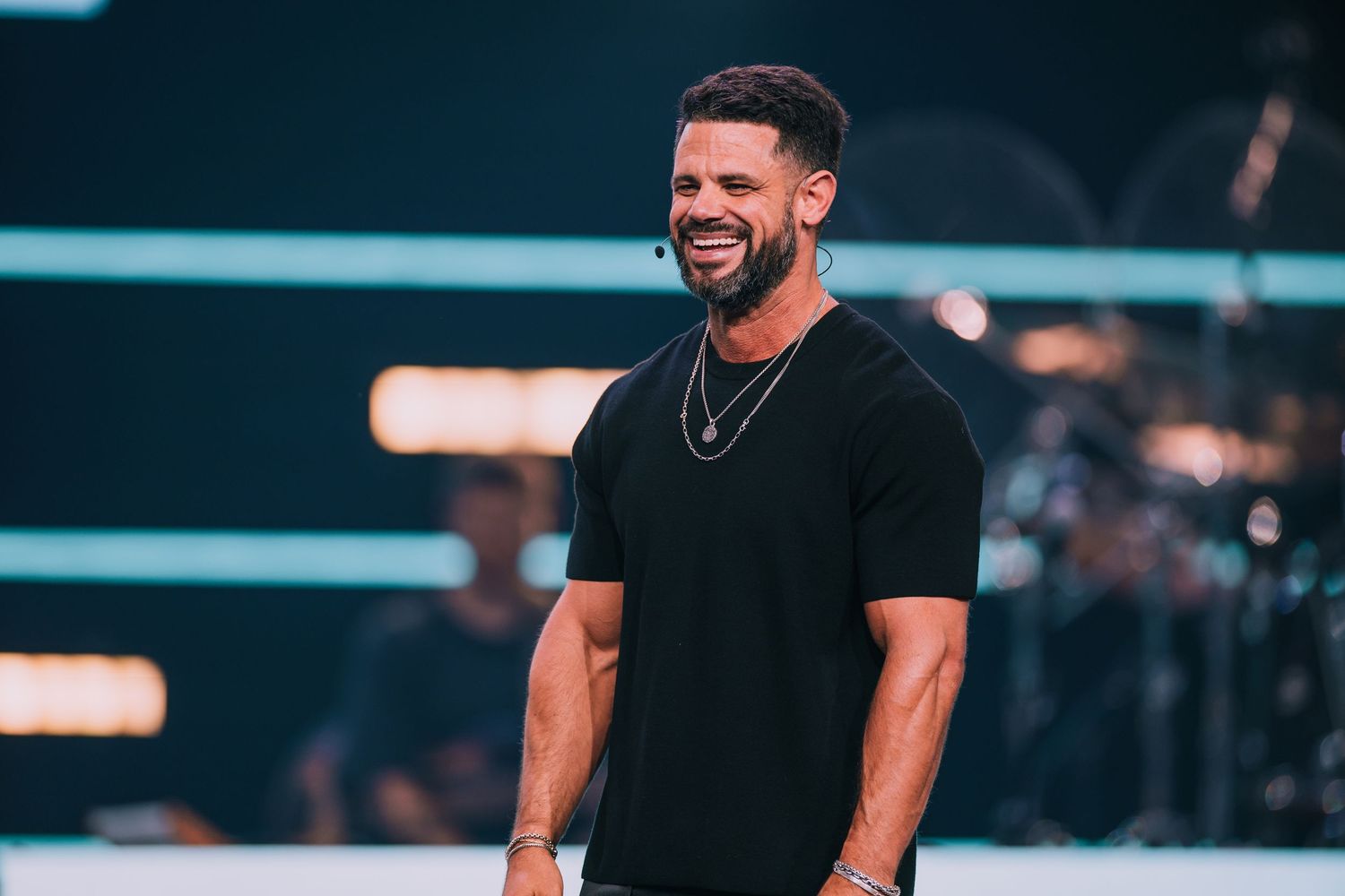 Athletic man in black t-shirt smiling broadly on a church stage.