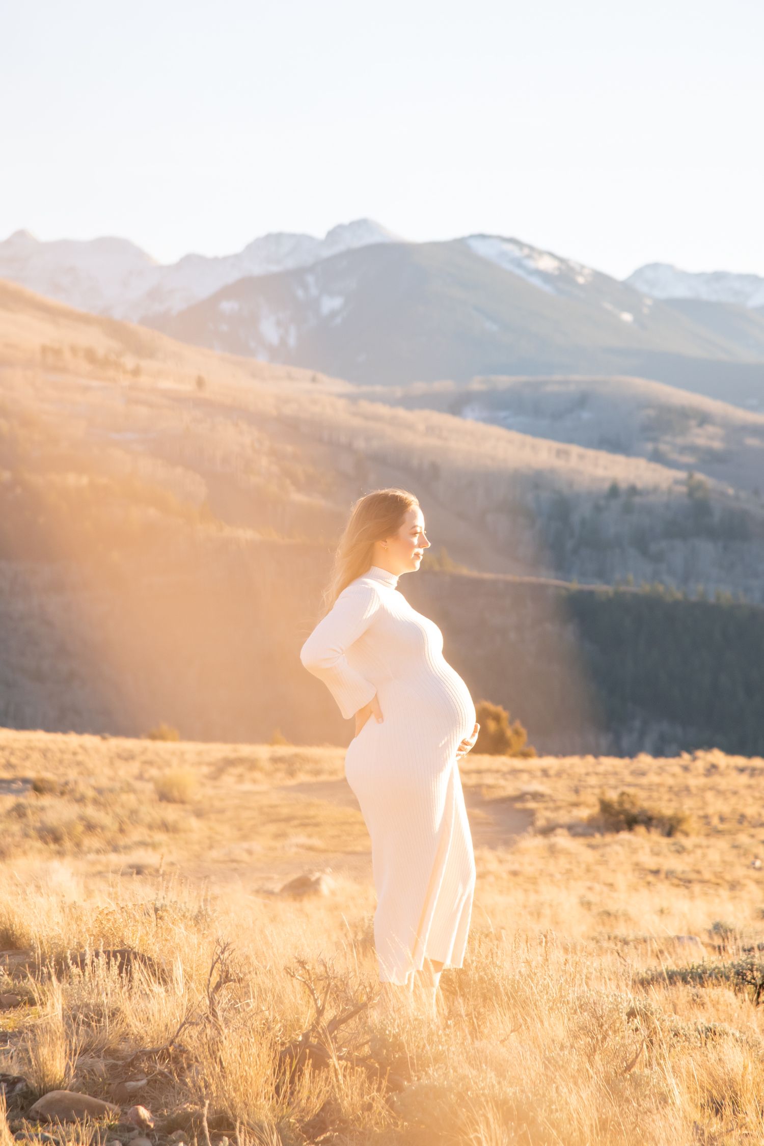 A pregnant woman in a white dress stands on a hilltop at golden hour with mountains in the background staring into the distance