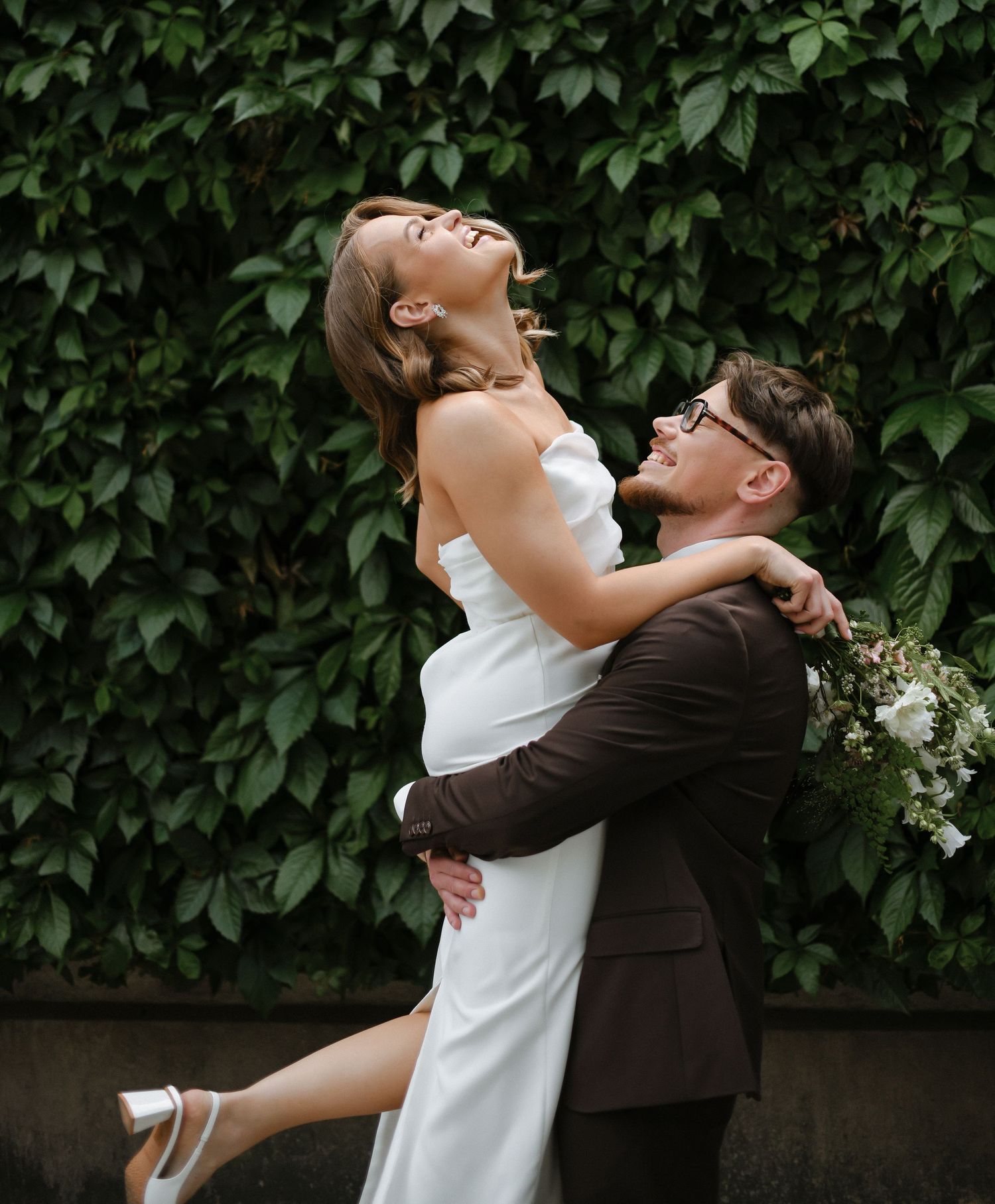 A couple in wedding attire embraces and laughs joyfully against a backdrop of lush green foliage.