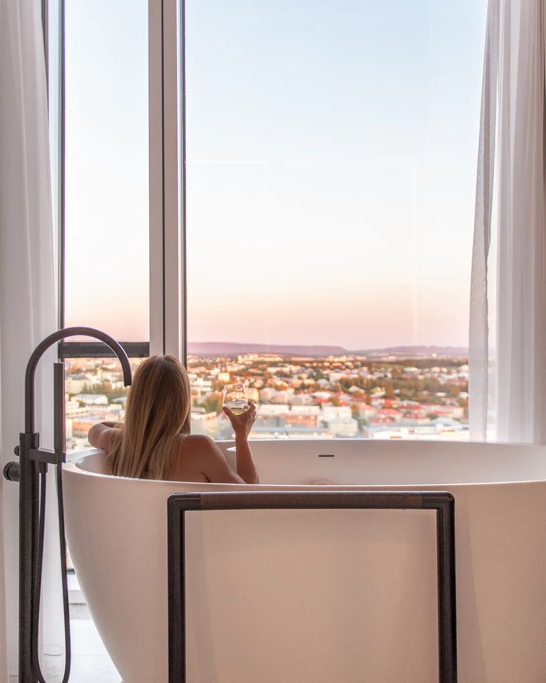 Person sitting in a bathtub looking out a large window over a city view.