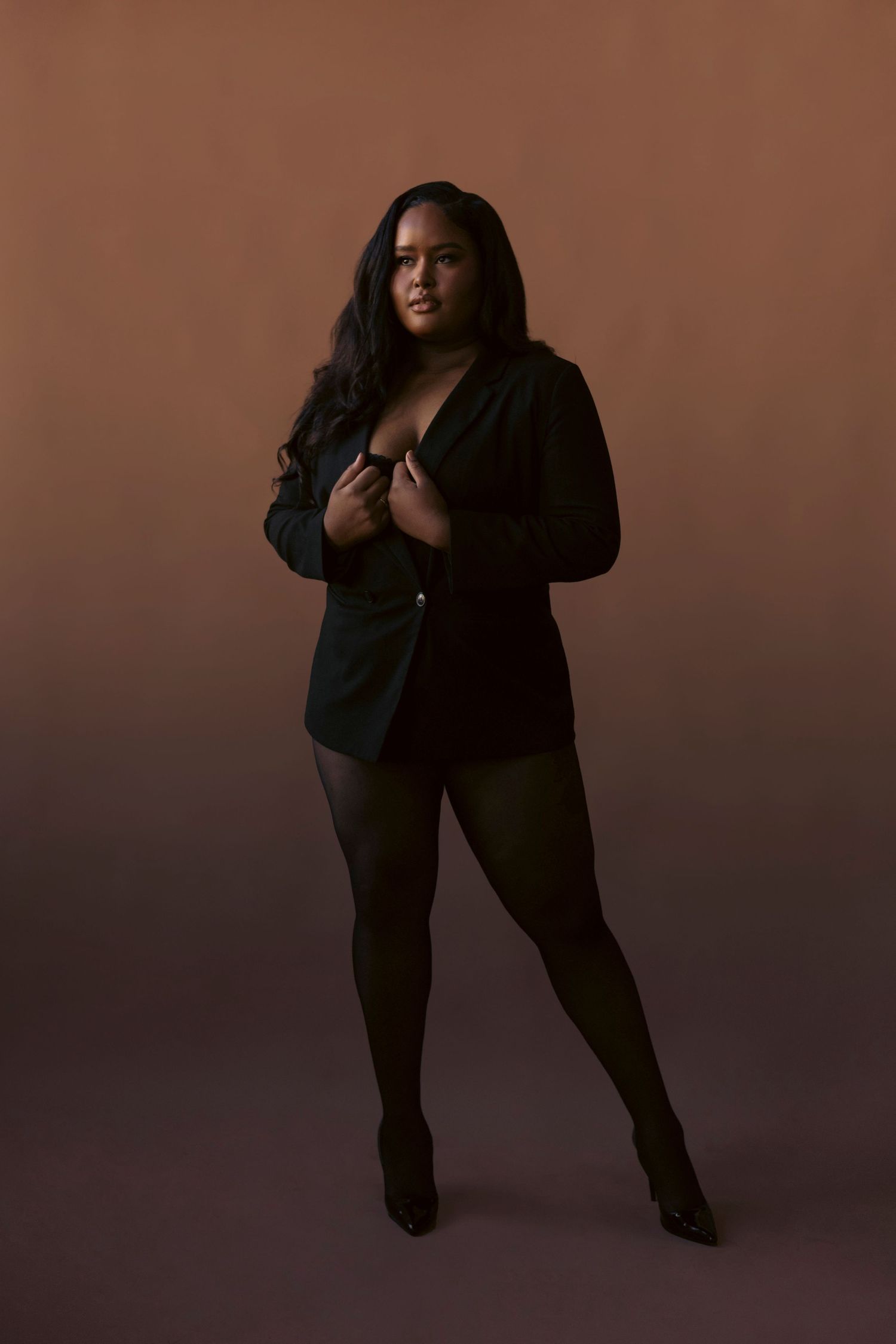 A curvy model in a black blazer and tights poses confidently against a brown studio backdrop.