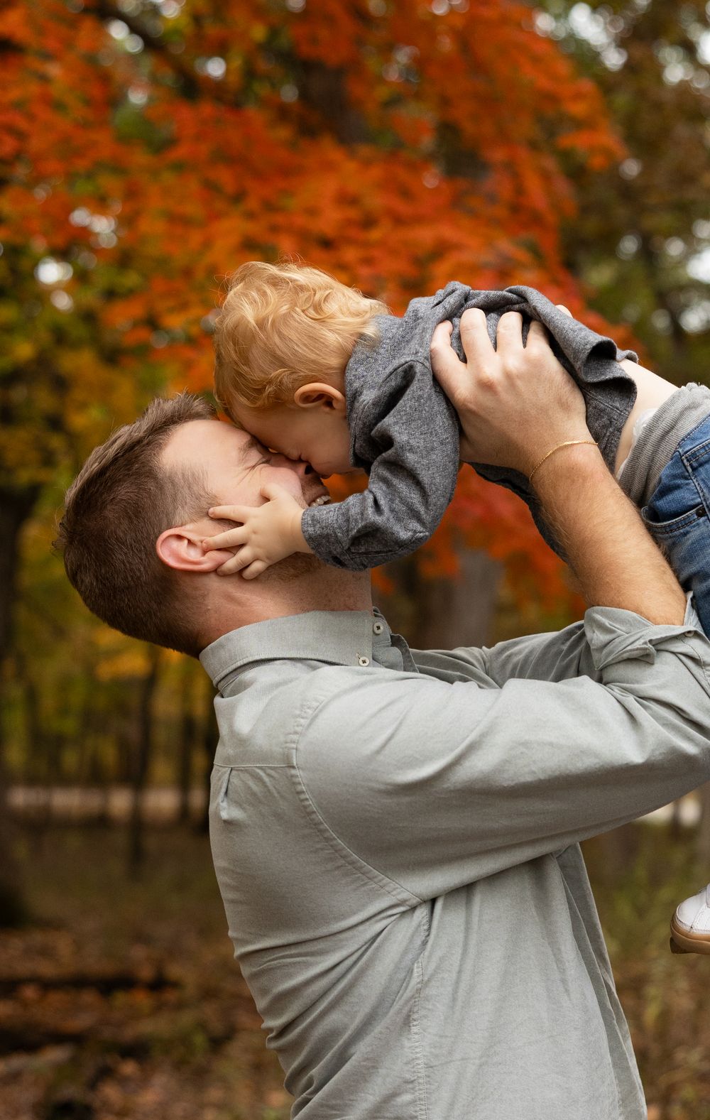 Father lifting toddler for a kiss among vibrant autumn foliage.