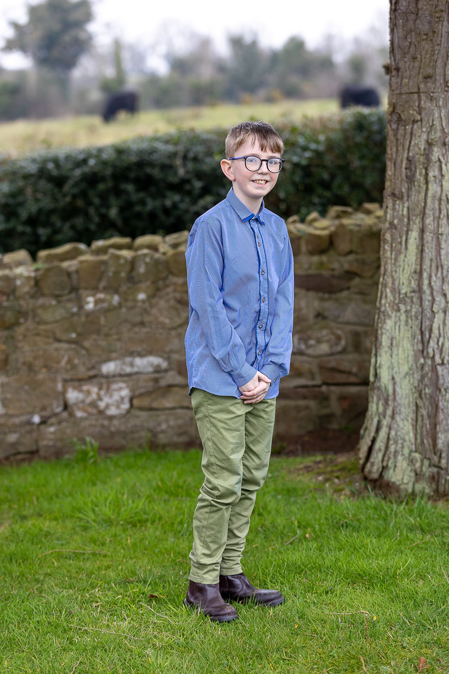 A young person in a blue shirt and khaki pants poses playfully on grass near a stone wall.