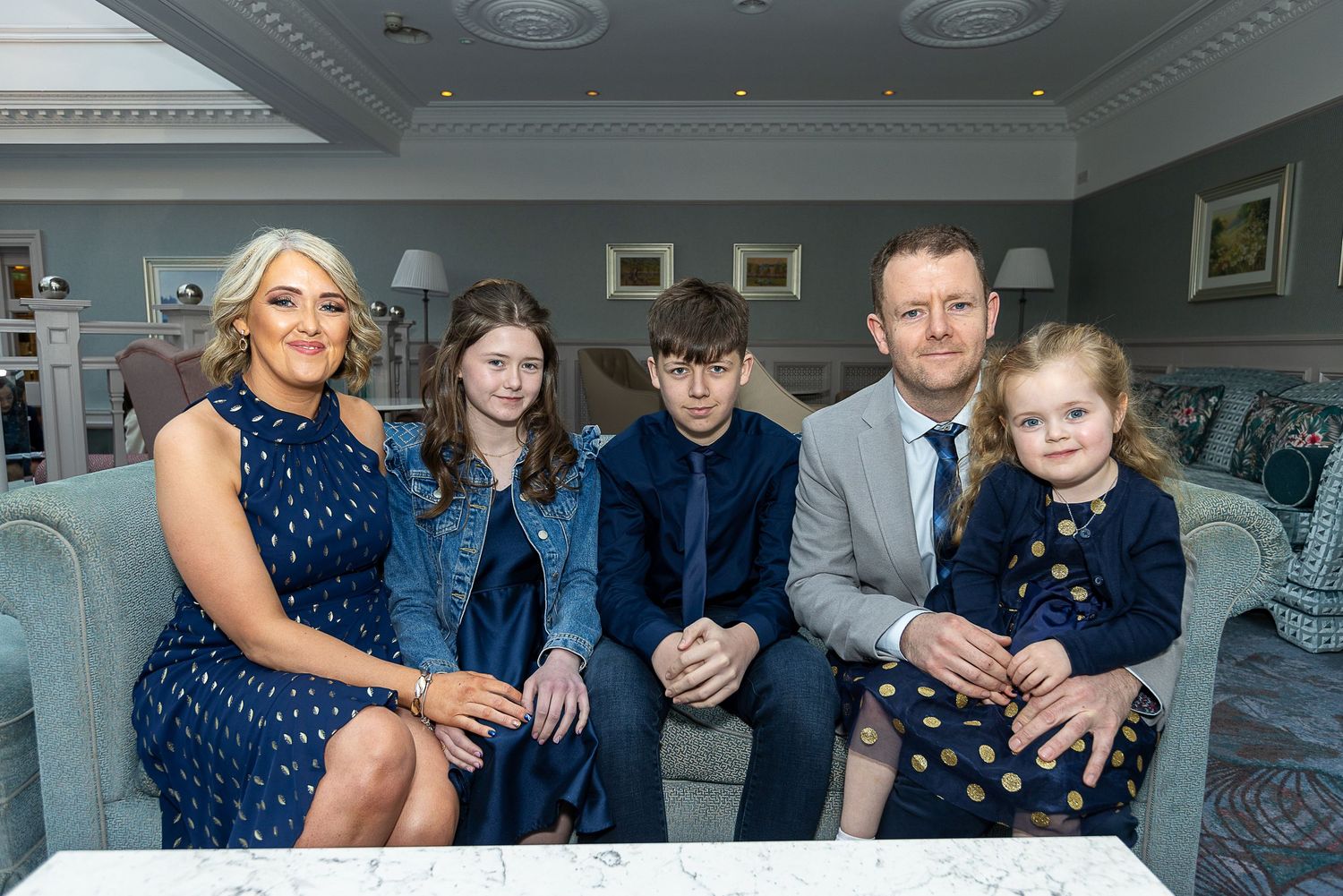 A family group sits together on an elegant blue sofa in a formal living room setting.