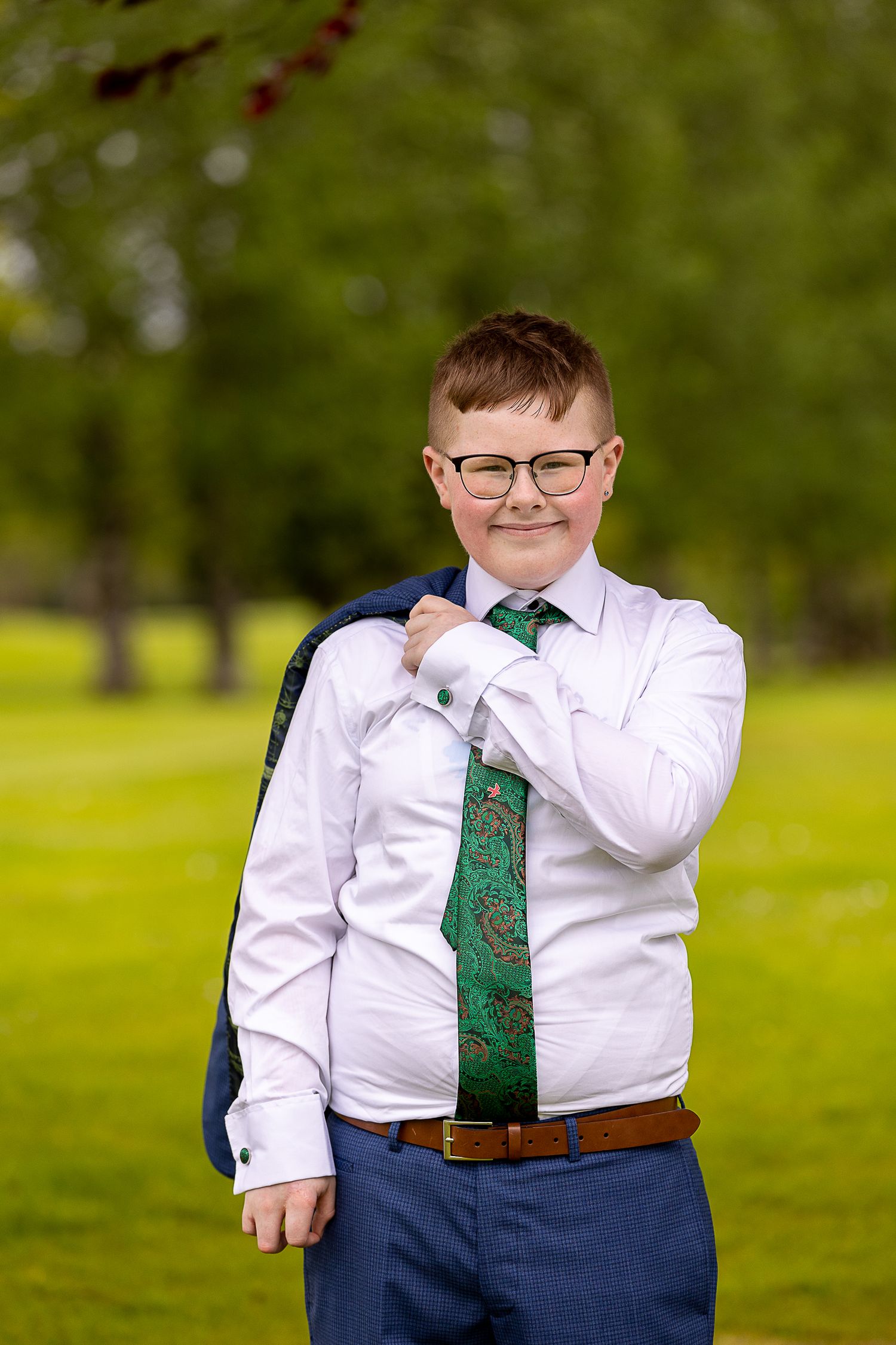A person wearing a white dress shirt and green tie poses outdoors in a grassy field on a sunny day.