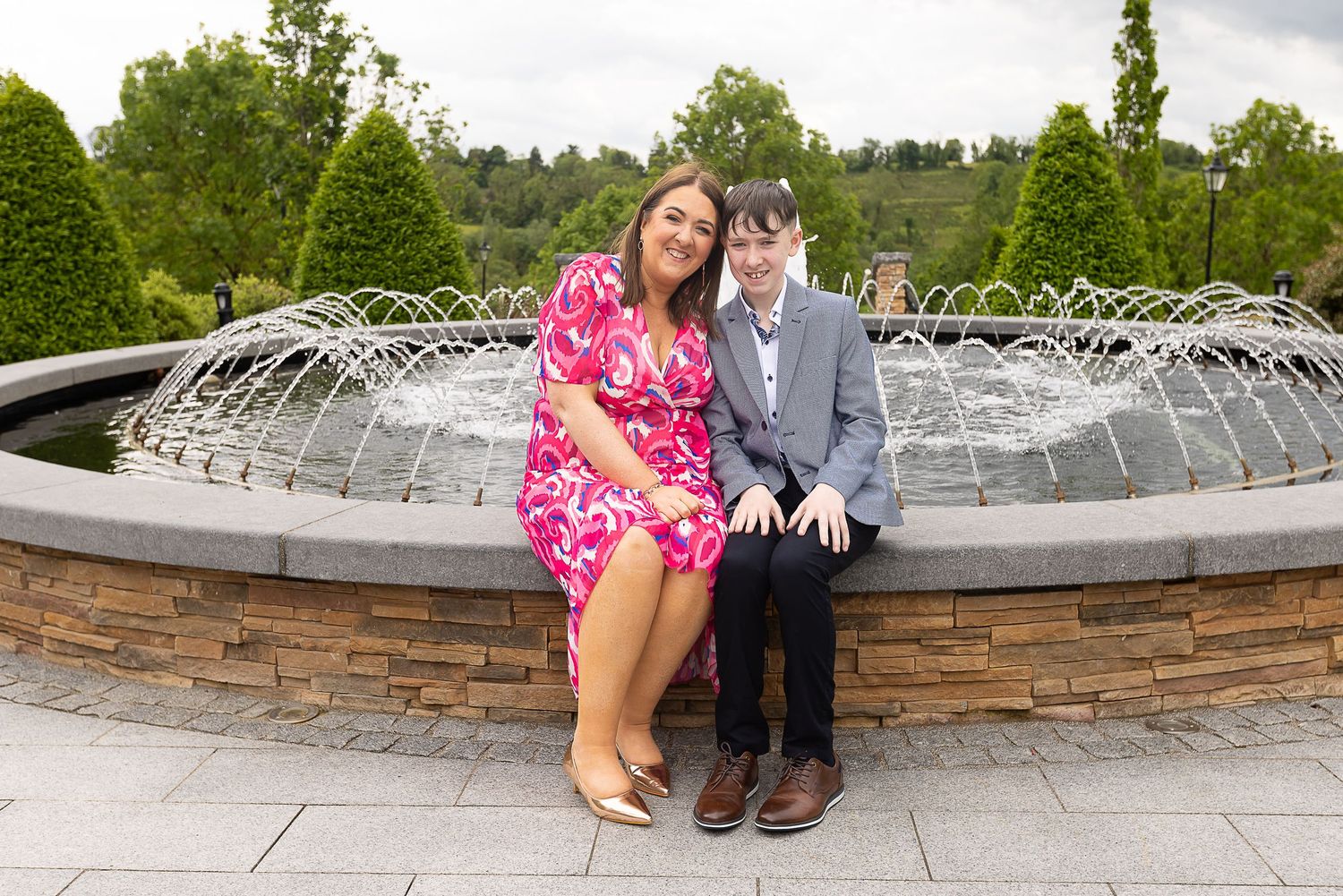 A couple sits together at the edge of a stone fountain in a garden setting.