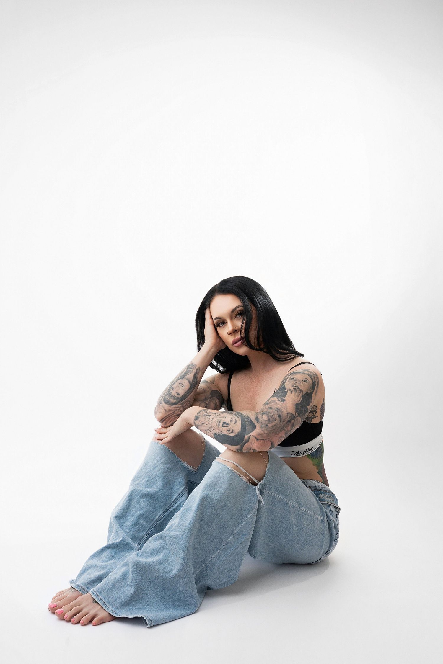 Artistic boudoir photo series showing tattooed figure in light denim jeans against white backdrop.