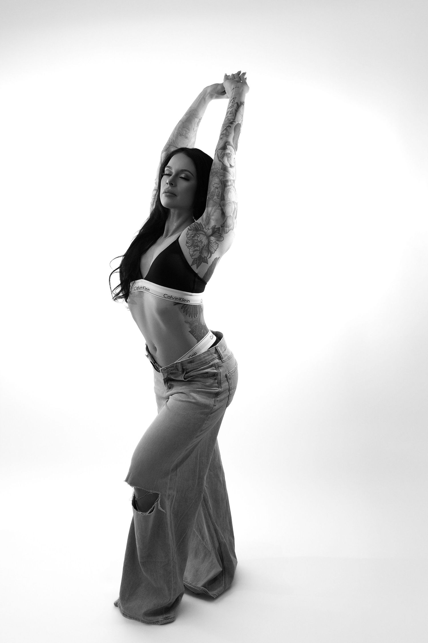 Black and white artistic dance pose with raised arms in casual jeans and crop top against white background.