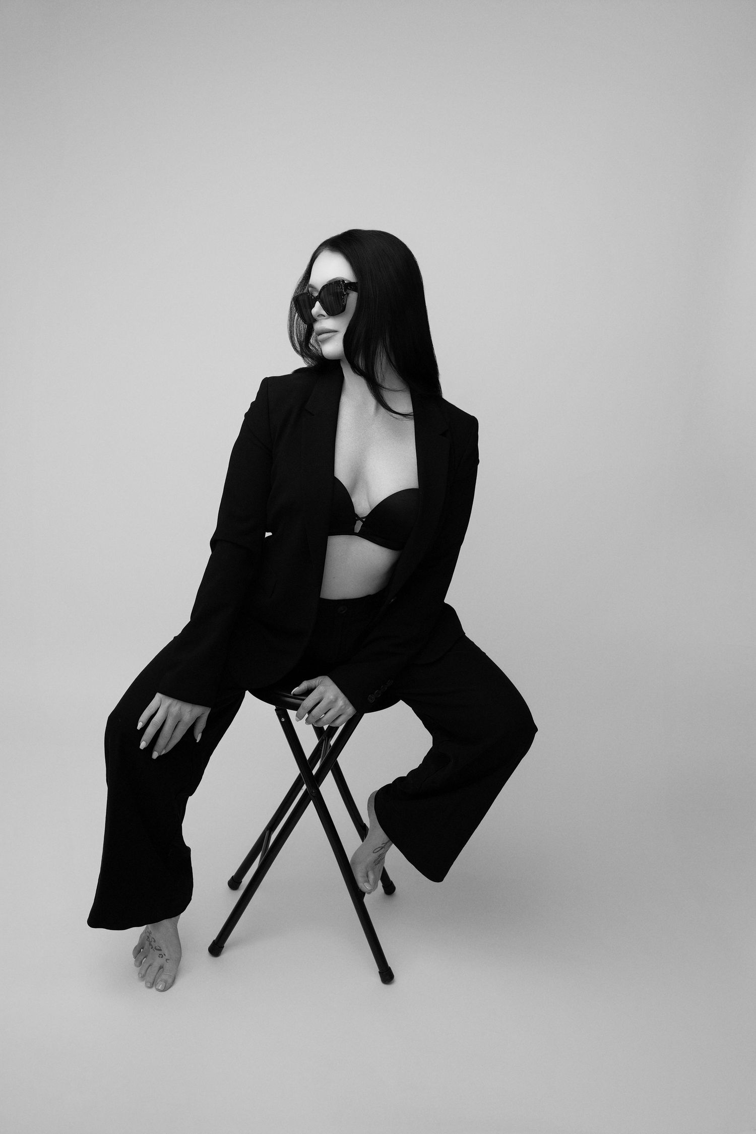 Artistic black and white fashion photo of a model in a black suit and sunglasses sitting on a stool against white backdrop.