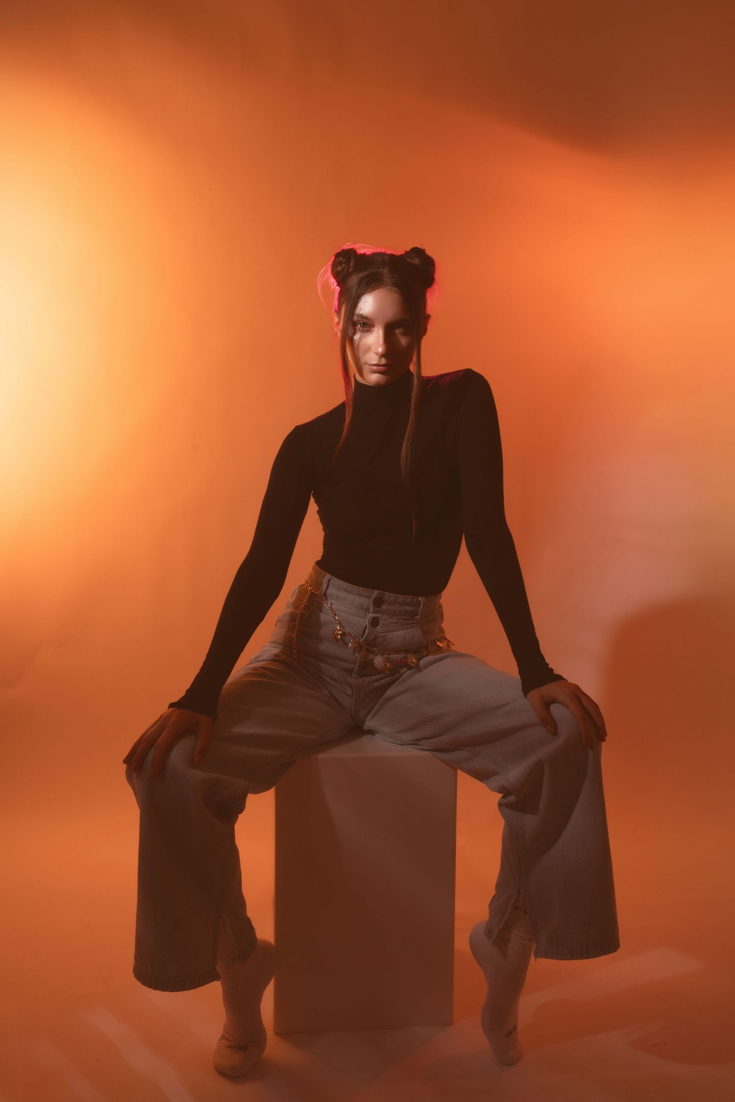 A silhouetted figure sits on a cube against an orange-tinted background in an artistic fashion photograph.