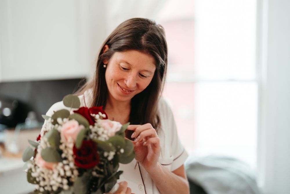 Woman in white sweater smelling fresh flowers in a bright, sunlit room with a dreamy expression.