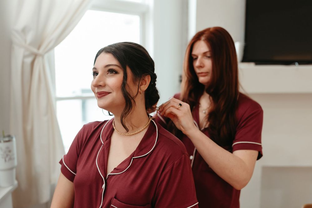 A bride getting her hair styled while wearing a burgundy robe in a bright sunlit room.