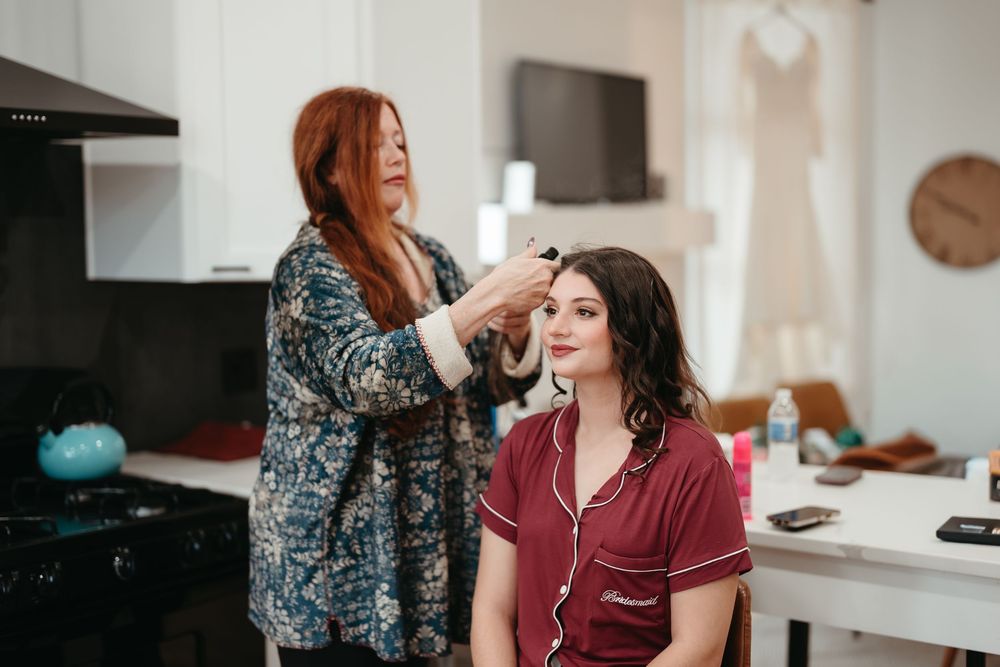 A stylist applies makeup to a client wearing a burgundy robe in a bright kitchen setting.