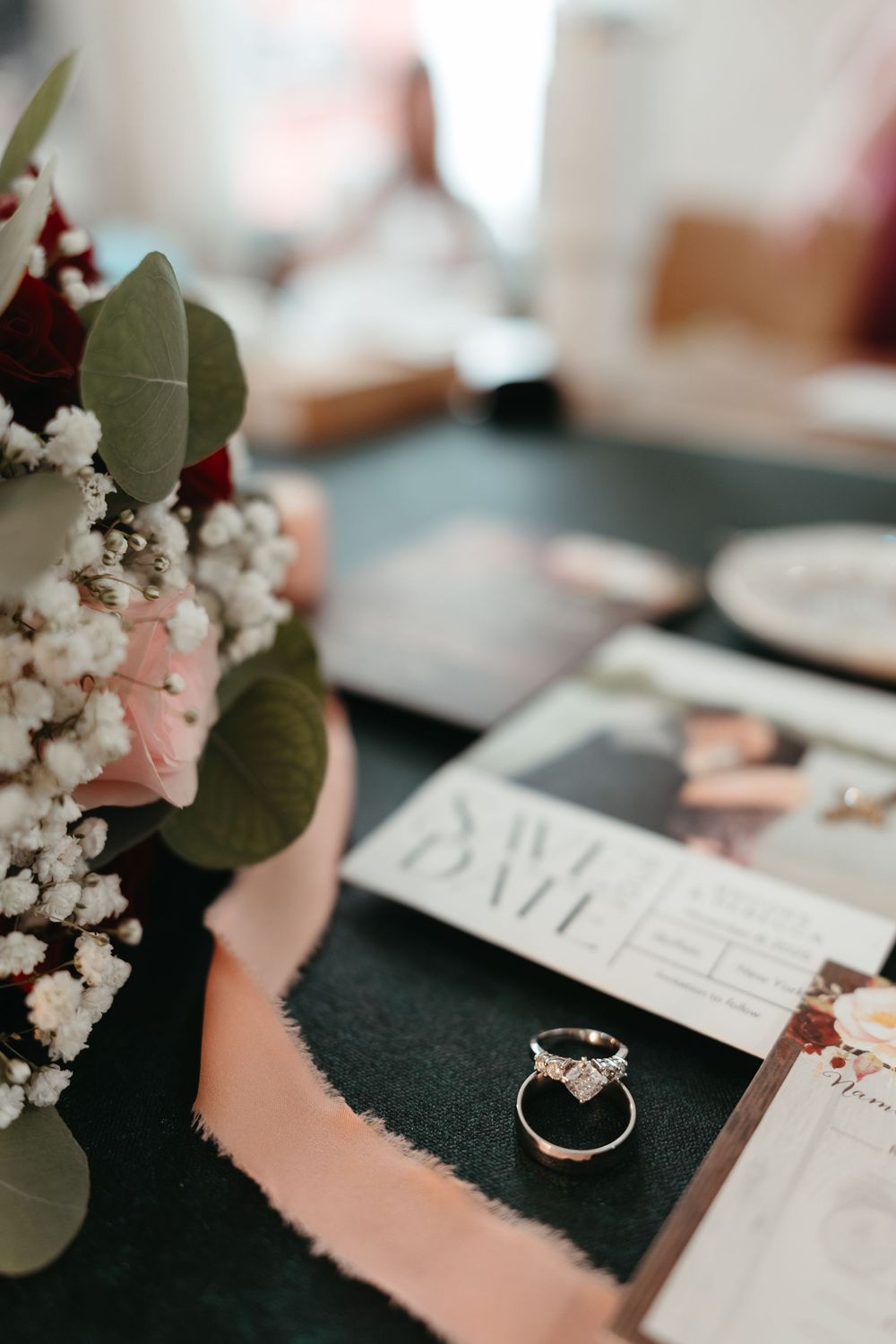 Wedding rings and floral arrangements with baby's breath and eucalyptus displayed on dark surface with pink accents.