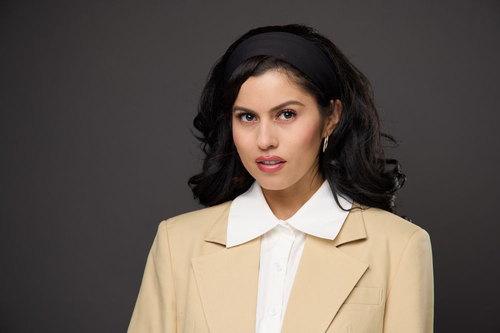 Professional headshot against dark background wearing beige blazer and white collared shirt with long dark hair.