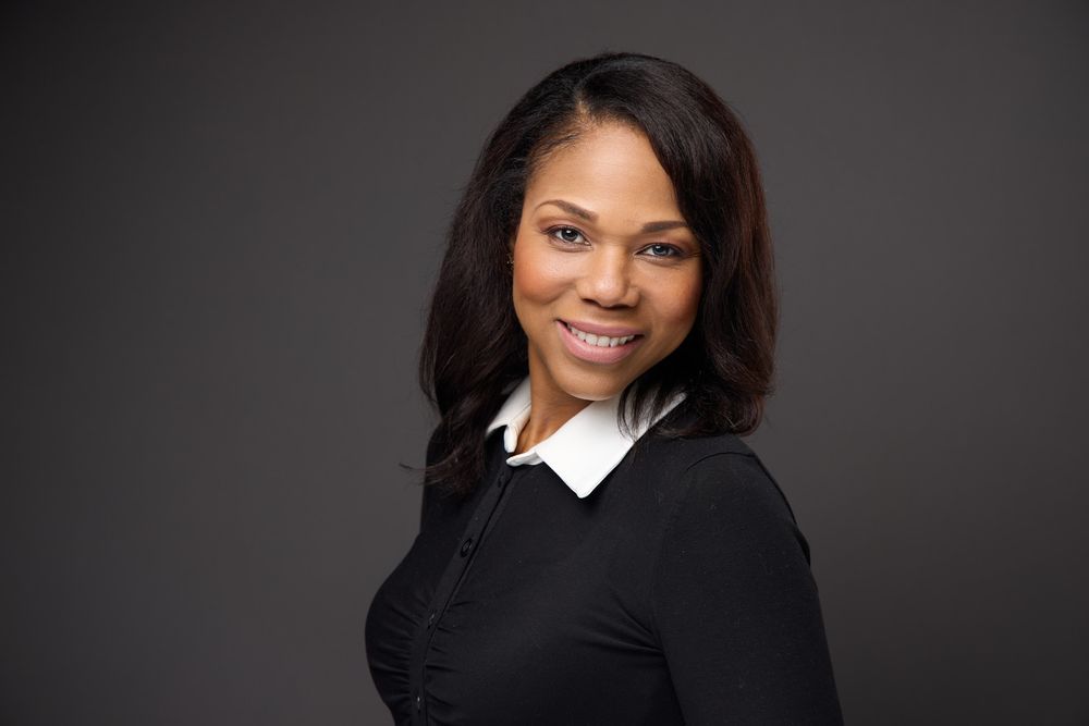 Professional headshot portrait of someone wearing a black sweater and white collar against a dark gray background.