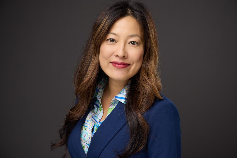 Professional headshot of a person wearing a navy blazer and patterned blouse against a dark background.