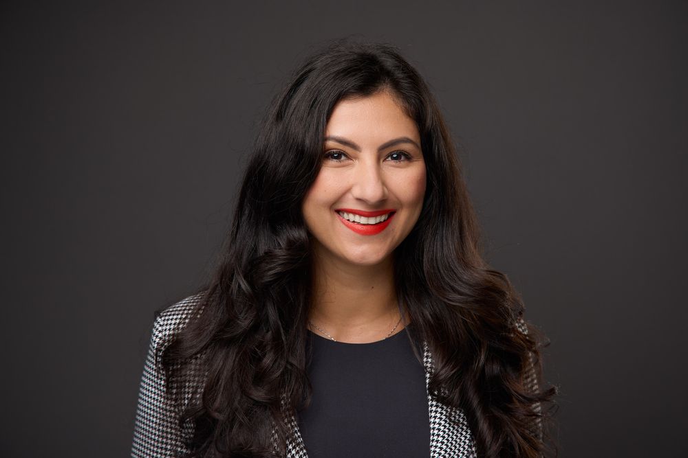 Professional headshot portrait of a woman with dark hair wearing a black top against a dark background.