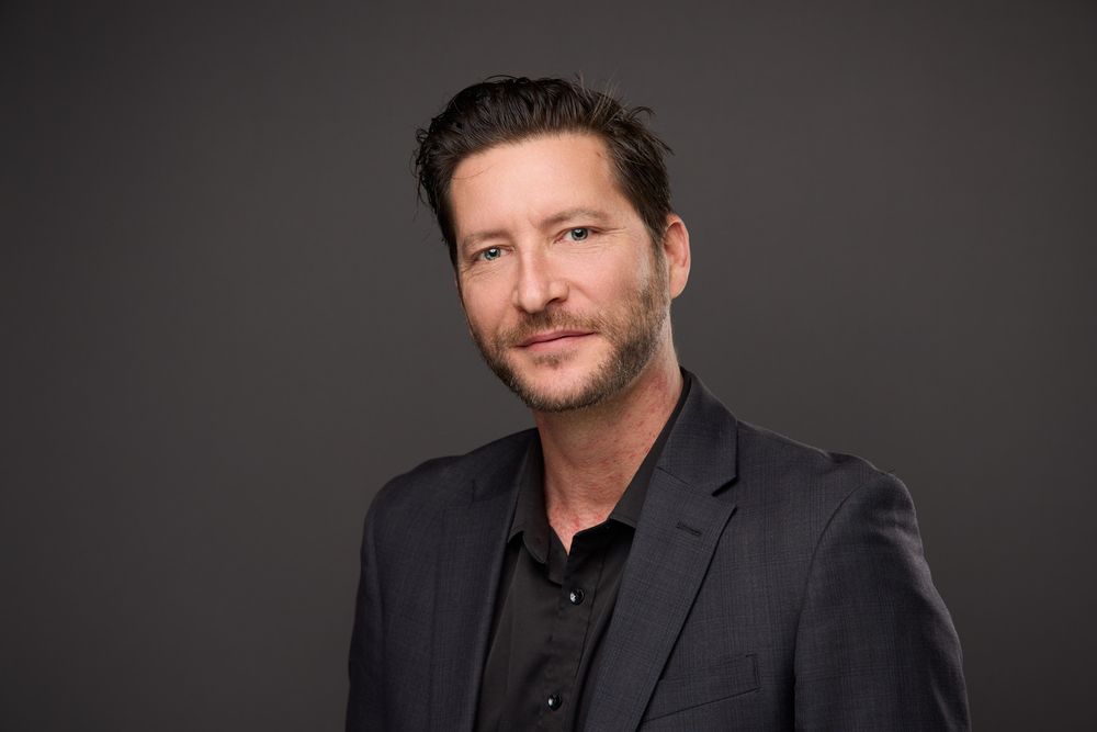 A person in a dark suit jacket and black shirt smiles while looking at the camera against a dark background.