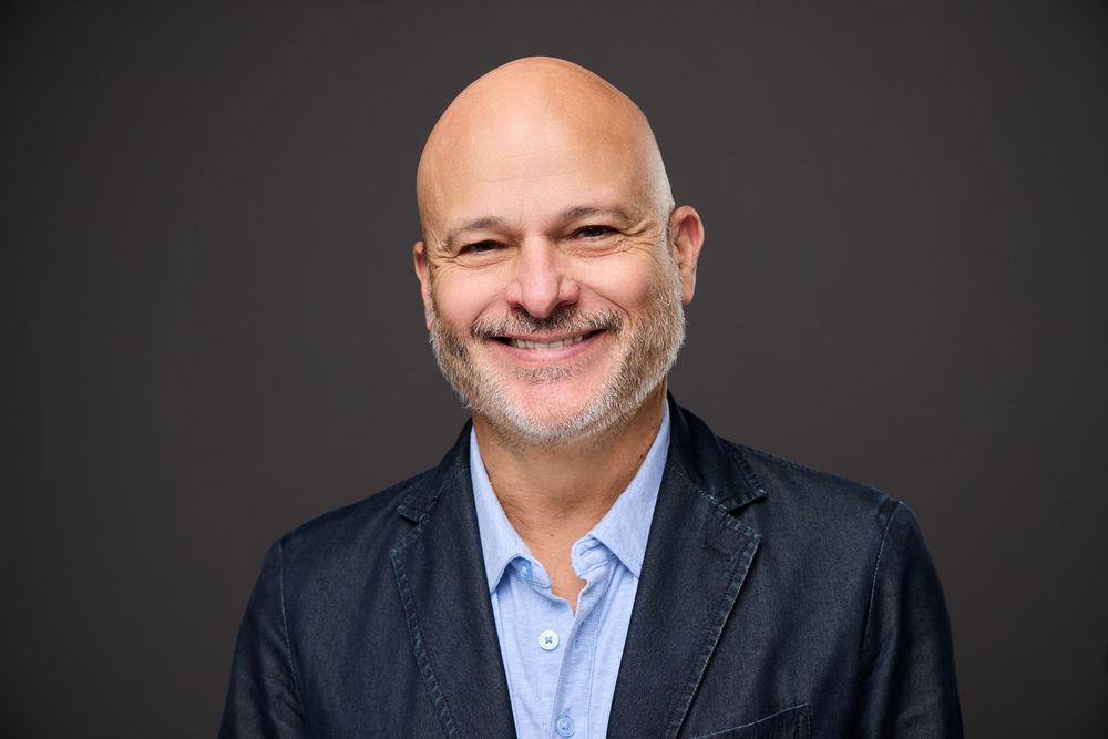 Professional headshot of someone in a dark suit and light blue dress shirt against a dark background.