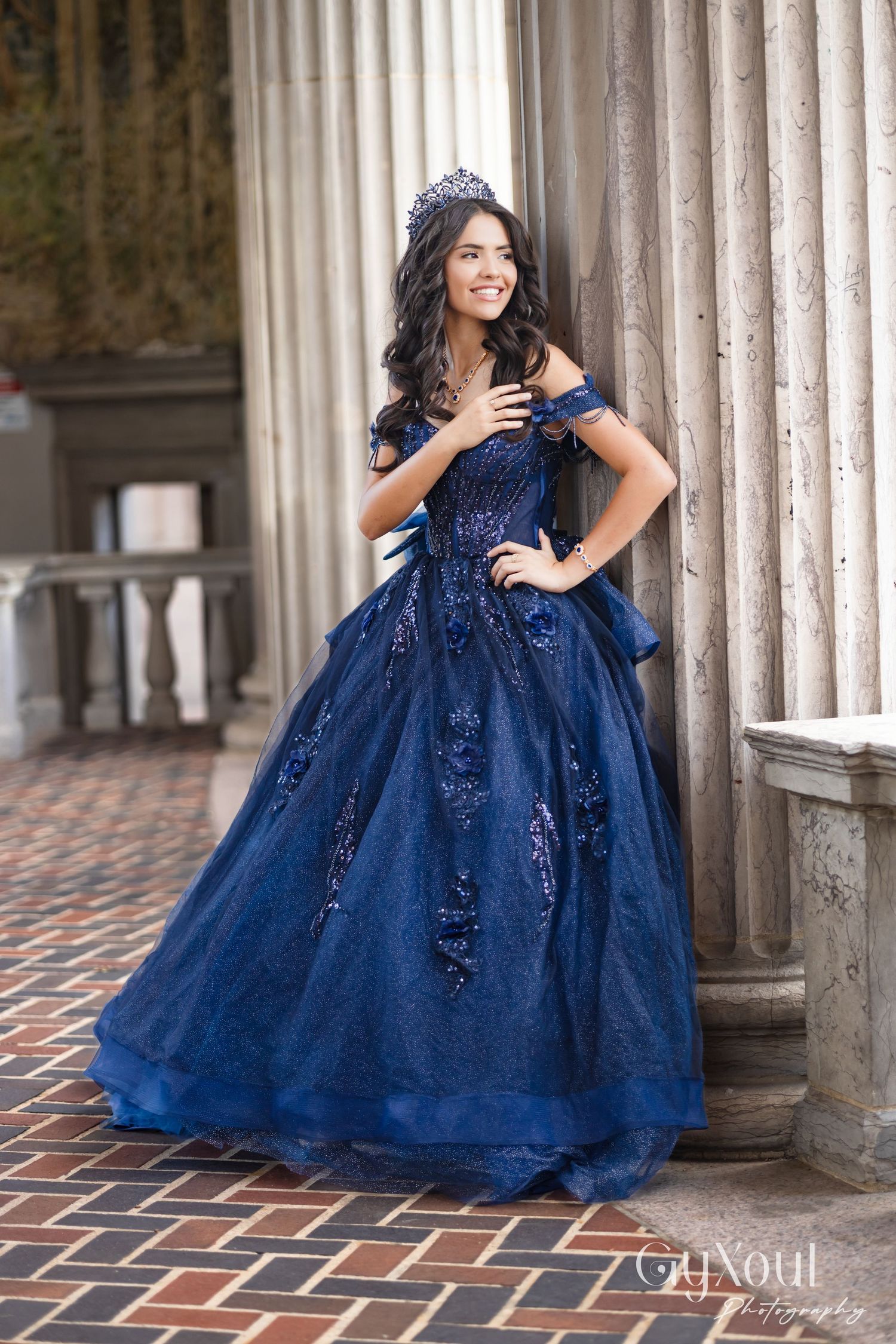 A young woman in a sparkling navy blue ball gown poses elegantly among classical stone columns and brick walkways.