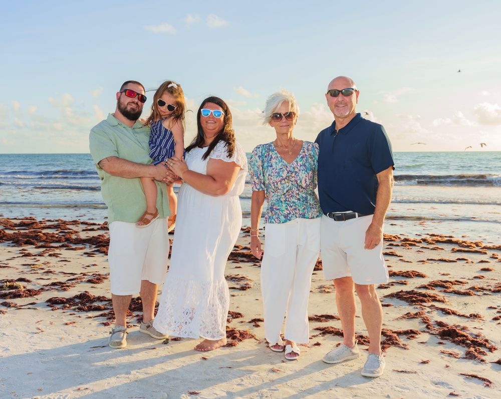 Family of five posing together on a sunny beach, wearing white bottoms and colorful tops, with calm ocean waves in the background.