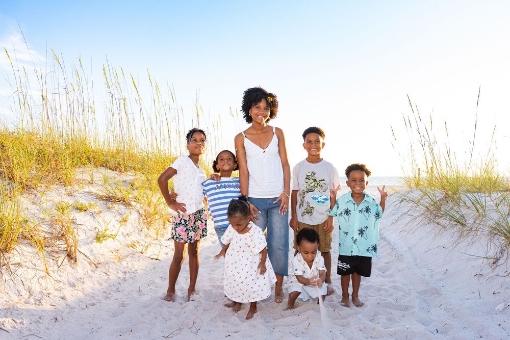 A woman and six children pose together on a sandy beach with sea grass and dunes in the background.