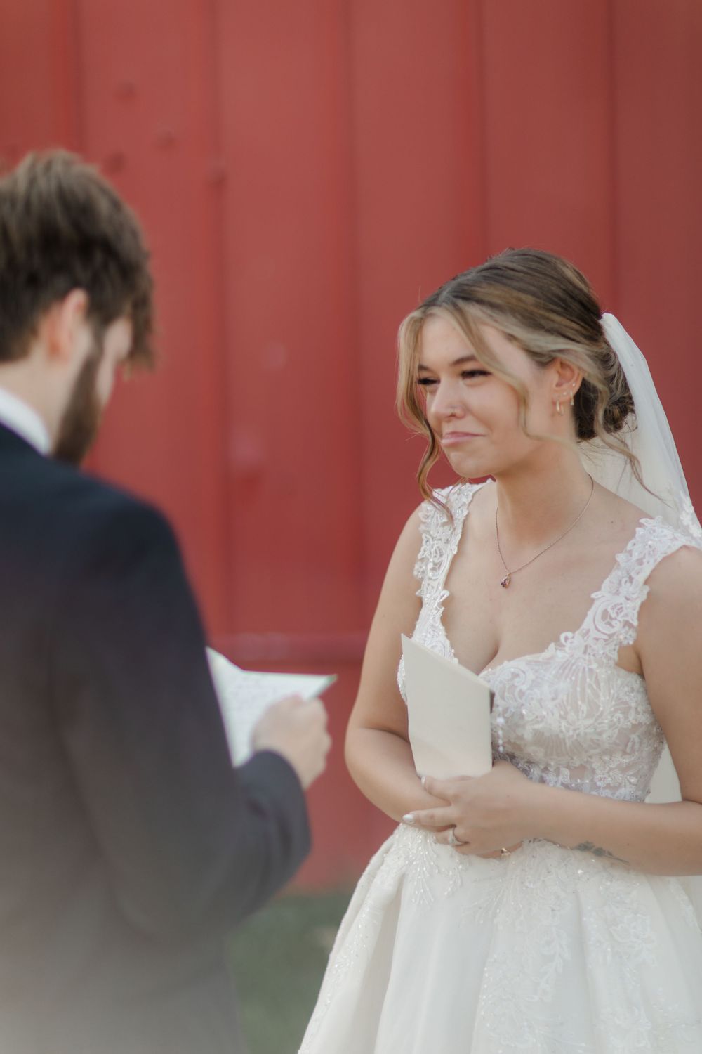 A bride in a white lace wedding dress shares a joyful moment while holding papers during an outdoor ceremony.