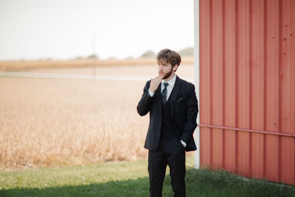 A couple in formal wedding attire poses together in front of a rustic red barn with golden wheat fields in the background.