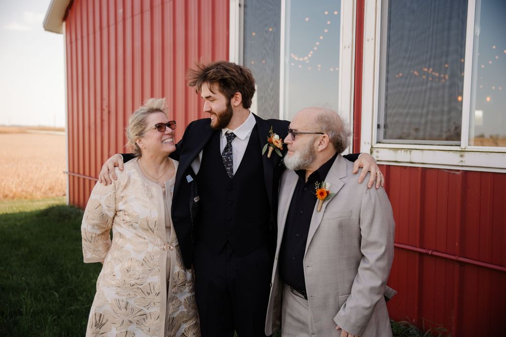 A wedding party poses together against a red barn wall in formal attire at sunset.