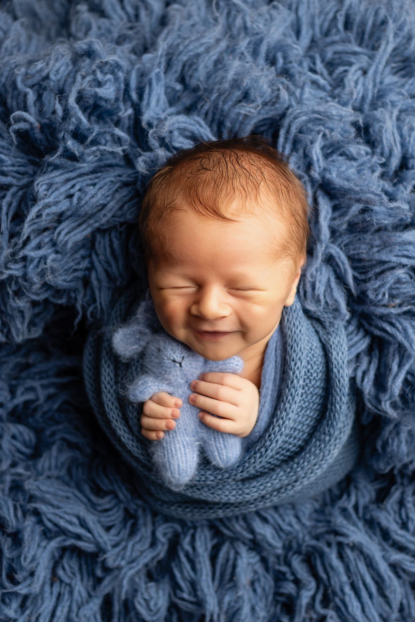 Newborn baby wrapped in cozy blue knit blanket smiling while sleeping on fluffy blue backdrop, taken by the best newborn photographer in atx