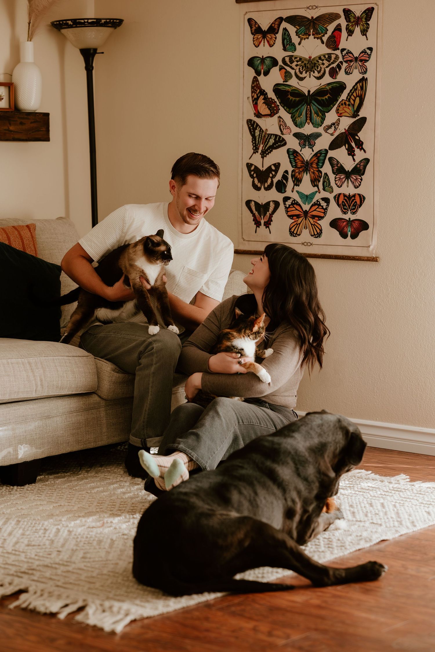 A couple relaxes on a hardwood floor with their pets beneath vintage butterfly artwork on the wall.