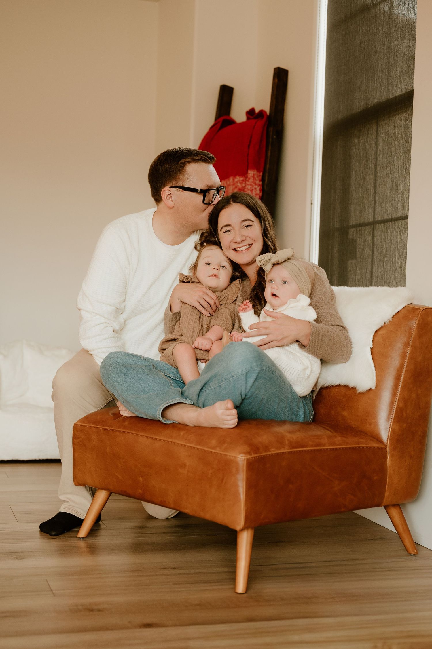 Family sits together on a brown leather chair in a cozy living room with natural lighting.