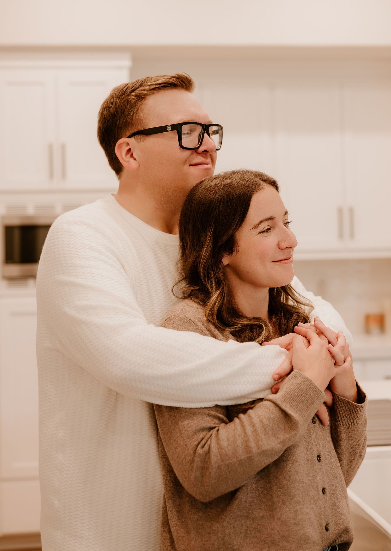 A couple in white and brown sweaters embraces tenderly in a bright kitchen setting.