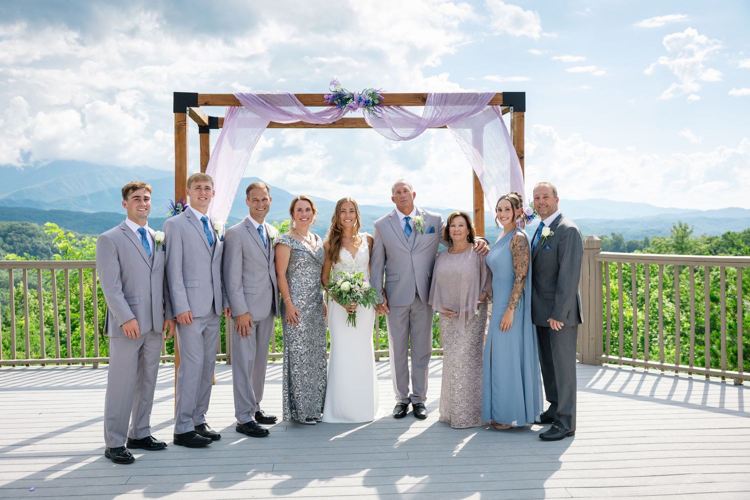 Wedding party in gray suits and light blue dresses posing together on an outdoor deck with mountain views.
