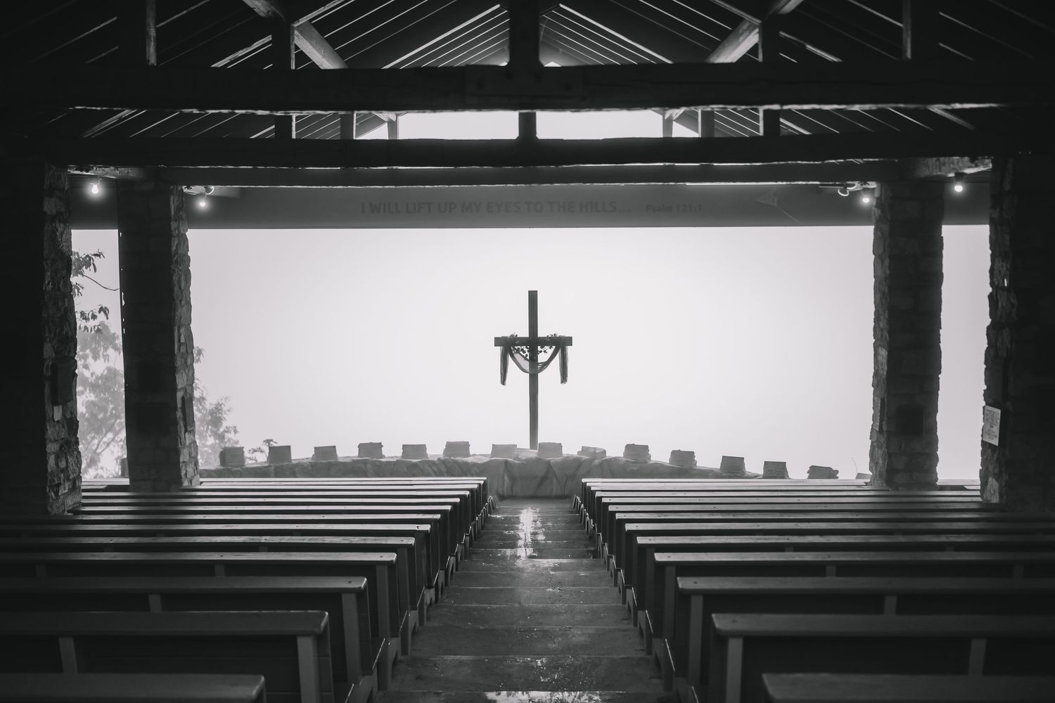 Black and white interior view of an empty chapel with wooden pews leading to a simple cross at the altar.