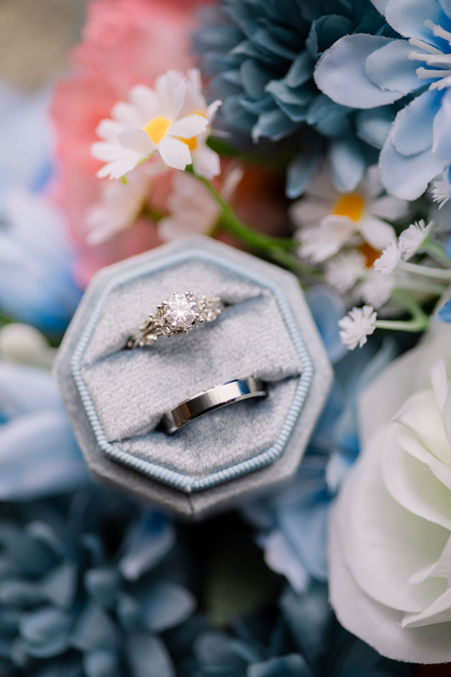 Wedding rings nestled in a light blue velvet octagonal ring box surrounded by blue and white flowers.