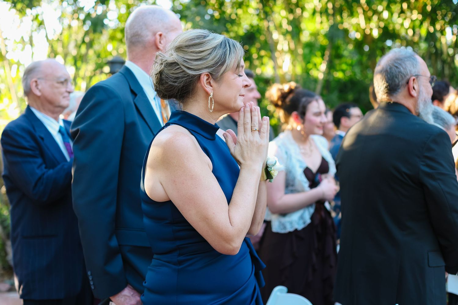 Wedding guests stand in a row outdoors during an emotional ceremony moment under green trees.