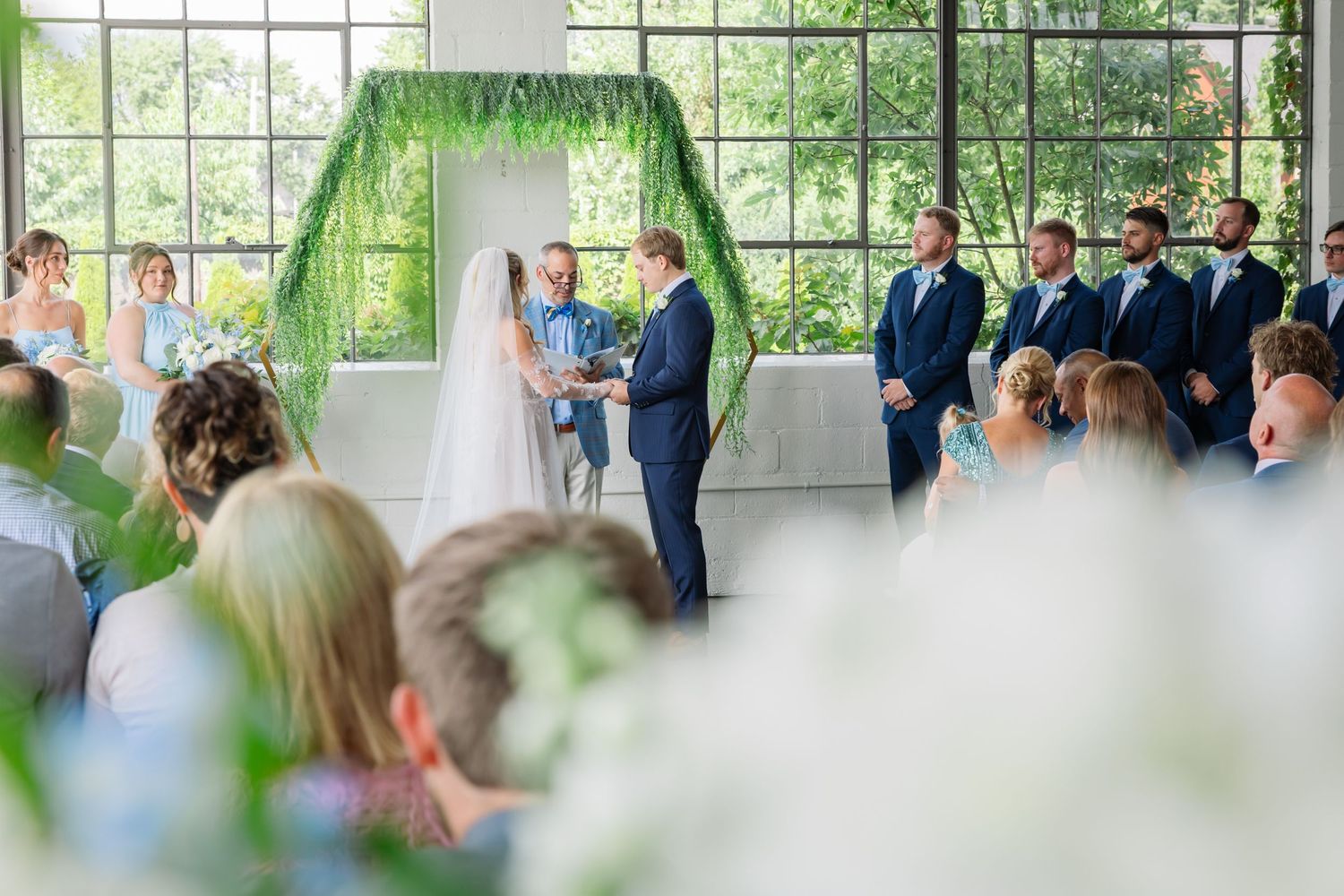 Wedding ceremony guests gather in a bright sunlit room with large windows and hanging greenery decorations.