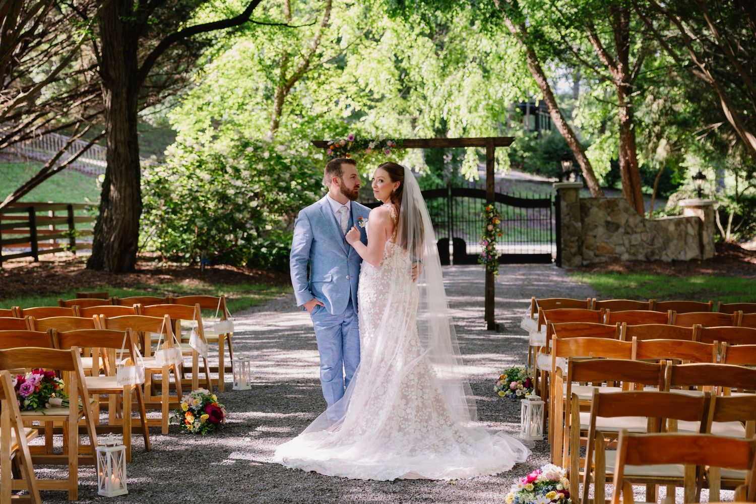 A romantic outdoor wedding ceremony setup with wooden chairs and a tree-lined path in a lush garden setting.