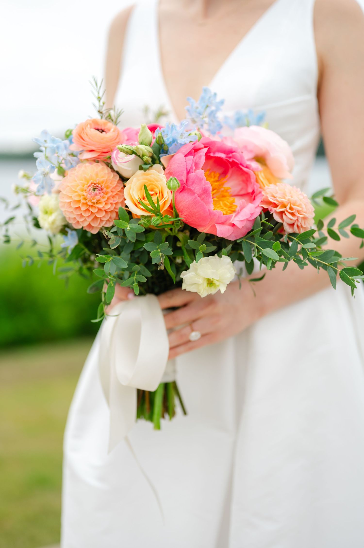 Newlywed couple embraces outdoors with vibrant coral and yellow floral bouquet against waterfront backdrop.