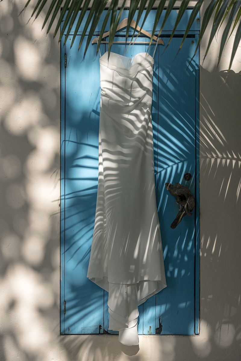 Wedding dress hanging on a blue door at Mahayana Tulum, illuminated by palm shadows; fine-art detail from a beachfront destination wedding.