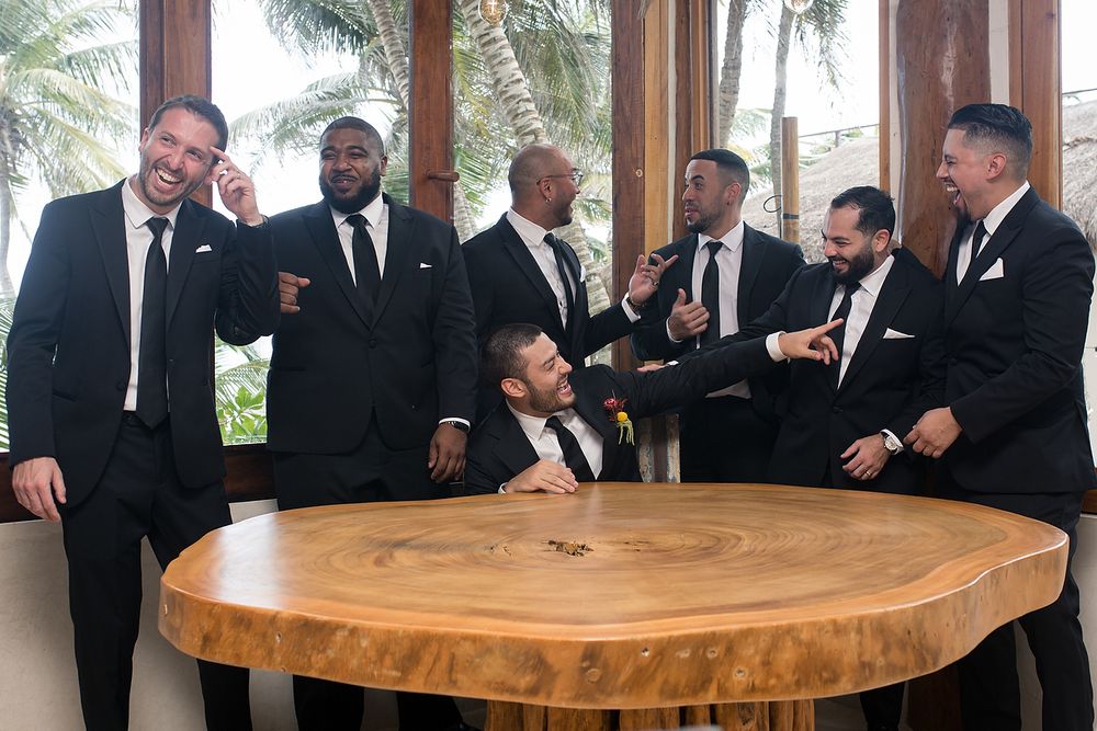 Groomsmen in black suits gather around a wooden table at an outdoor venue during a wedding celebration.