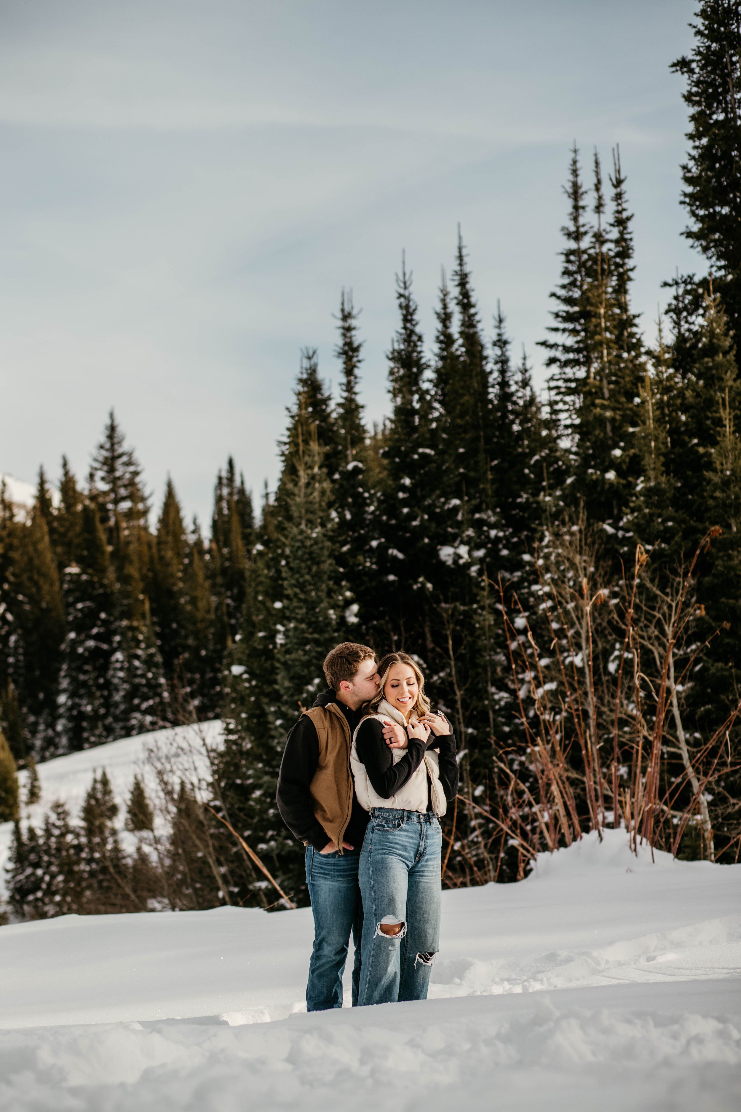 A couple shares an intimate moment in a snowy winter landscape surrounded by evergreen trees.