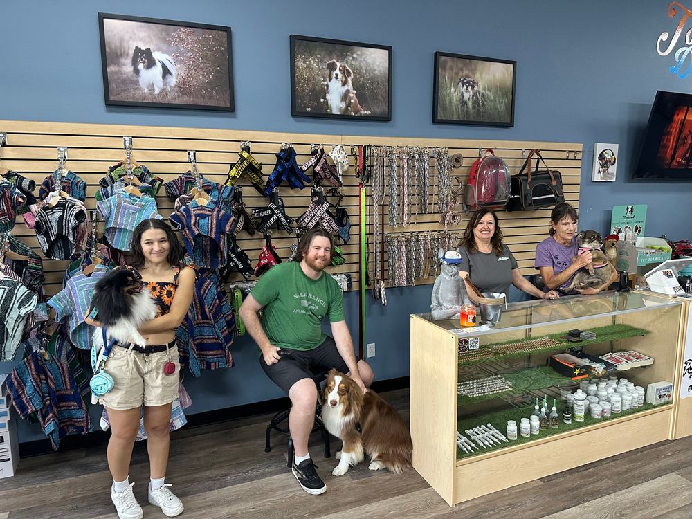 Pet store interior showing rows of supplies, merchandise displays, and customers browsing with their dogs.