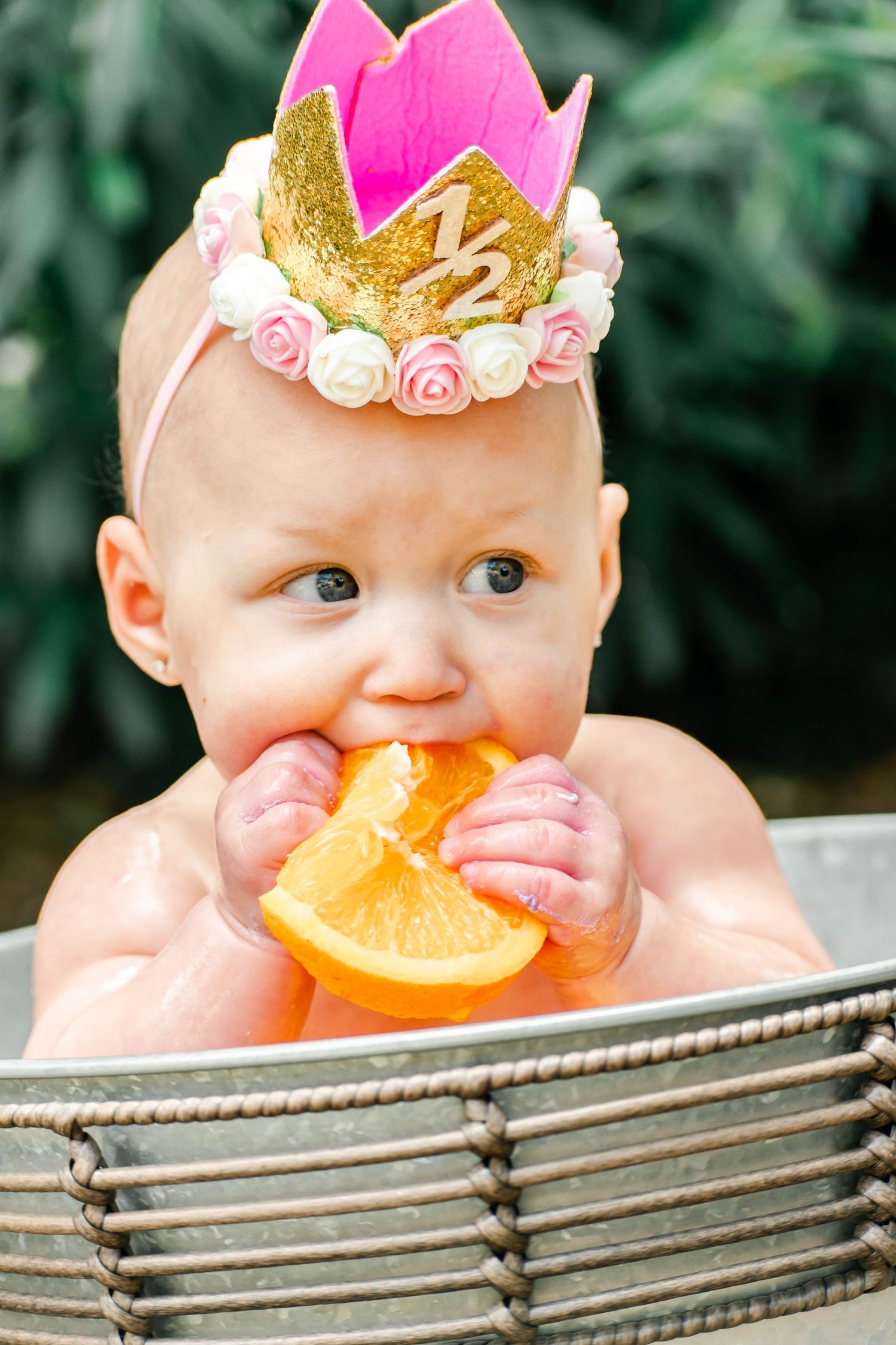 Baby wearing pink crown plays with orange slices while sitting in metal basin outdoors.
