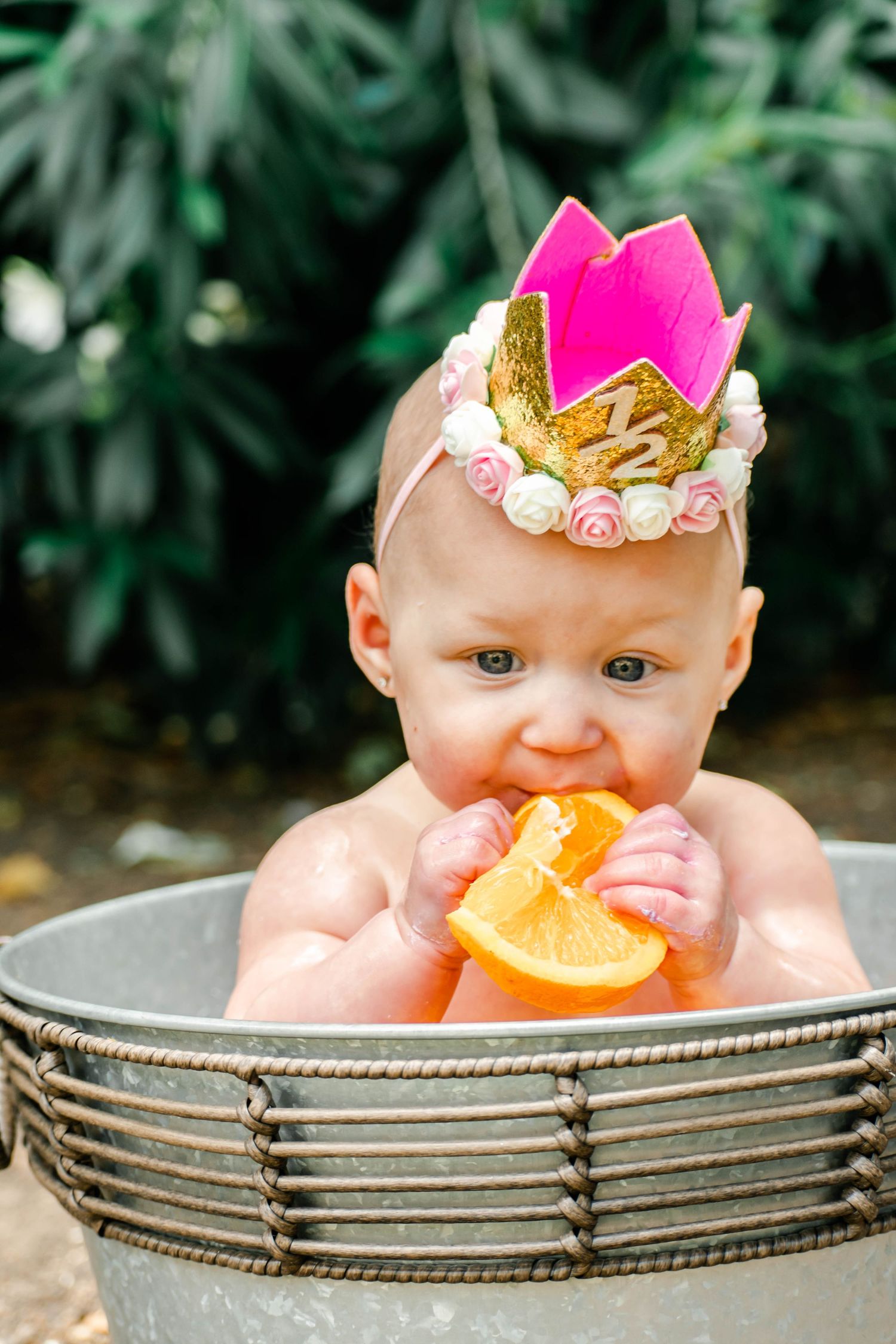 Small child in decorative pink crown tastes citrus fruit while sitting in rustic metal tub.